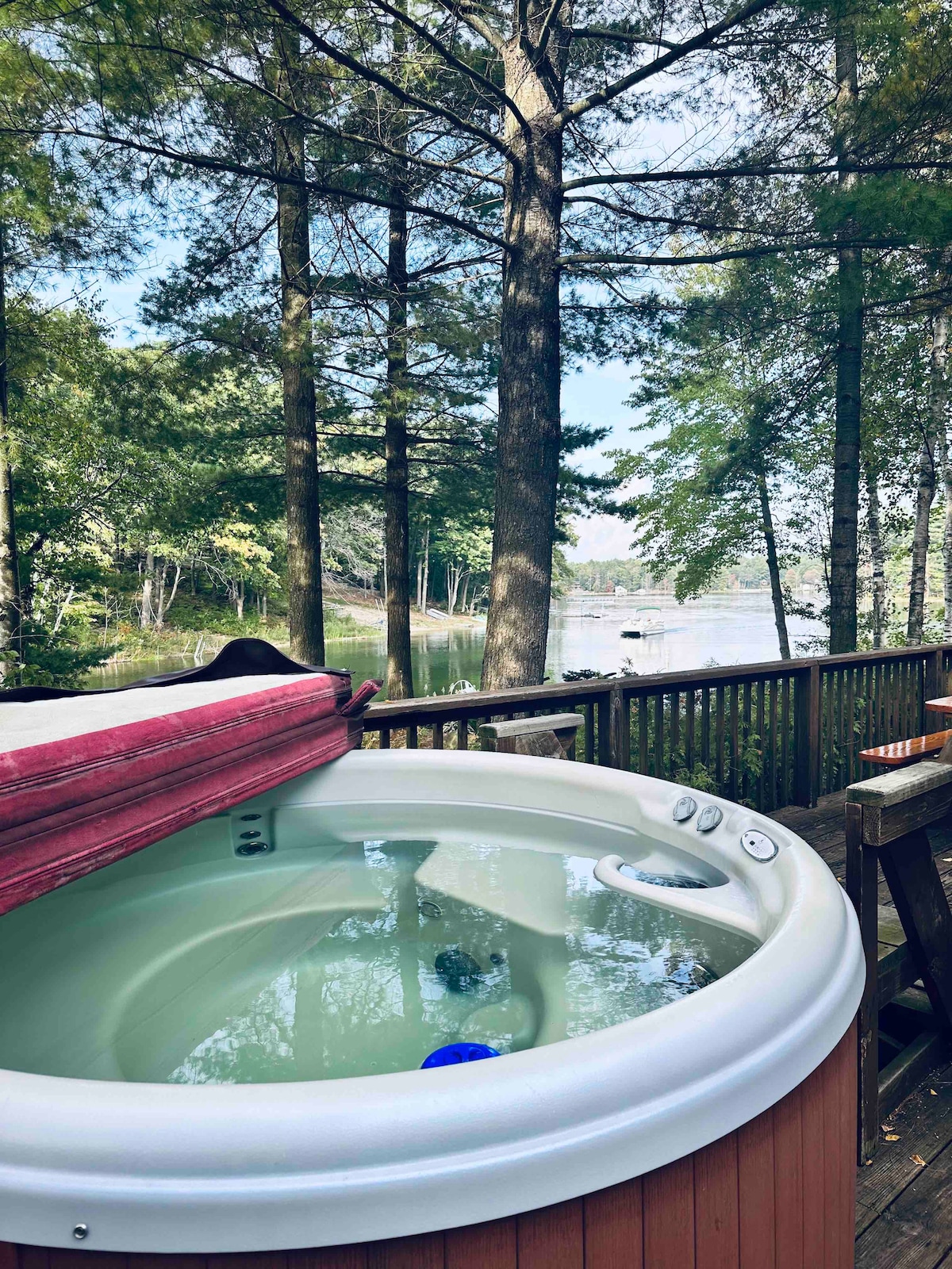 A hot tub is situated on a wooden deck, surrounded by tall trees that provide shade. The water appears calm and inviting, reflecting the greenery nearby. A peaceful lake view is visible in the background, enhancing the relaxation of the outdoor space.