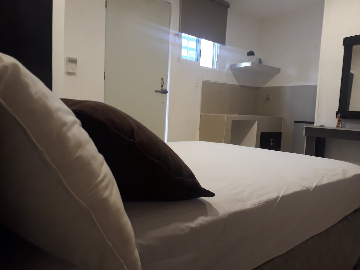 A comfortable sleeping area is shown, featuring a spacious bed with crisp white linens and a brown accent pillow. A simple bedside table is visible, along with a small kitchenette in the background. Natural light enters through a window, complementing the bright interior.