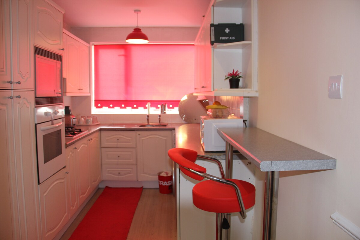 A tidy kitchen features white cabinetry and a red accent rug. A small breakfast bar with a chrome base and red stools is positioned beside a window with a red blind. Essential appliances, including a microwave and sink, are clearly visible.