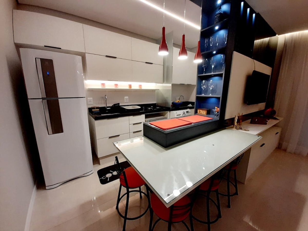 A modern kitchen space is equipped with sleek cabinetry and a large island featuring a glass top. Red chairs surround the island, while pendant lights hang above. A refrigerator and oven are integrated into the design, complemented by a stylish display shelf with glassware.