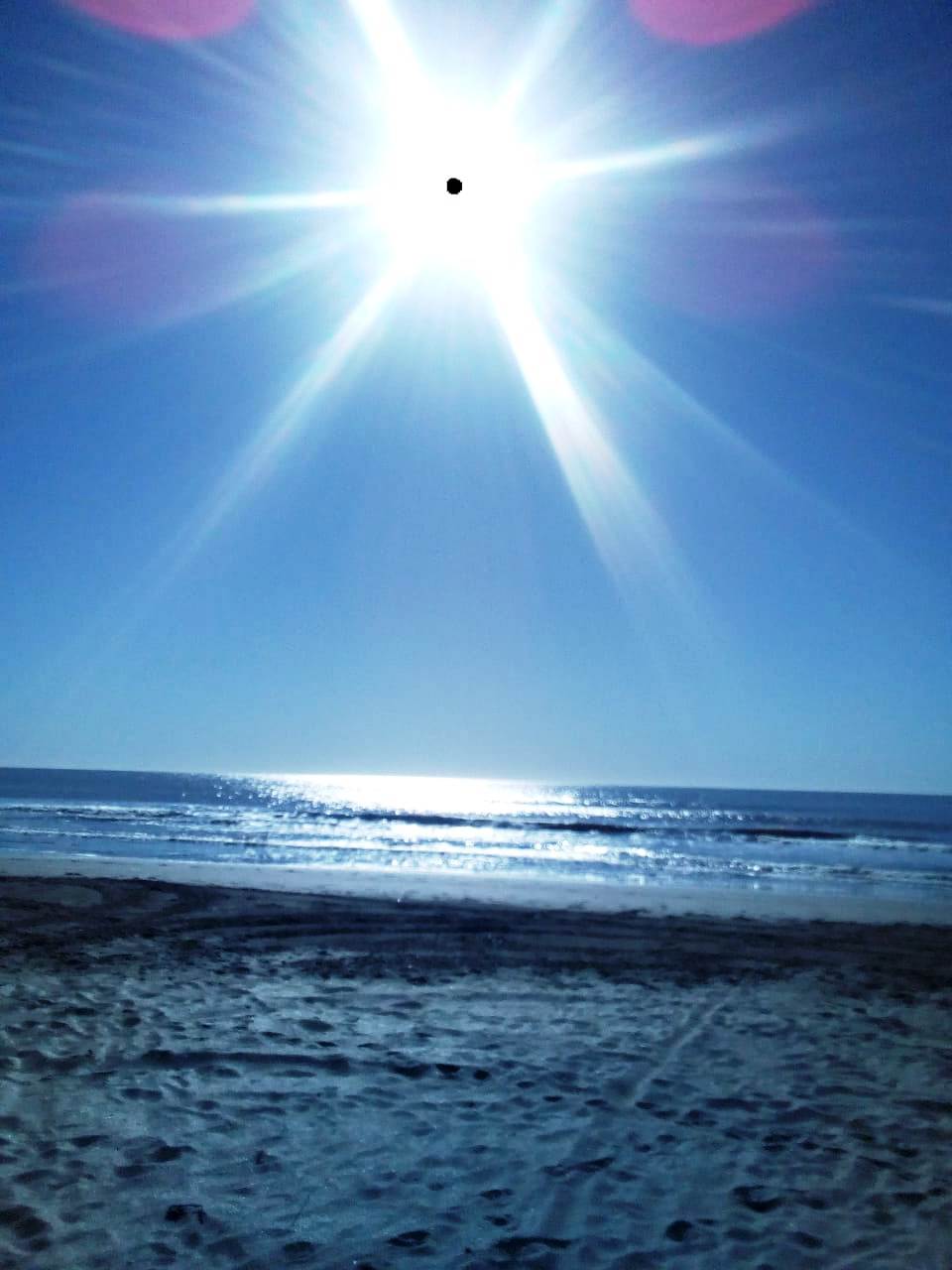 A bright sun is visible in a clear blue sky over a tranquil ocean. The sunlight reflects off the water's surface, creating a shimmering effect. The sandy beach is seen in the foreground, suggesting a serene coastal environment.