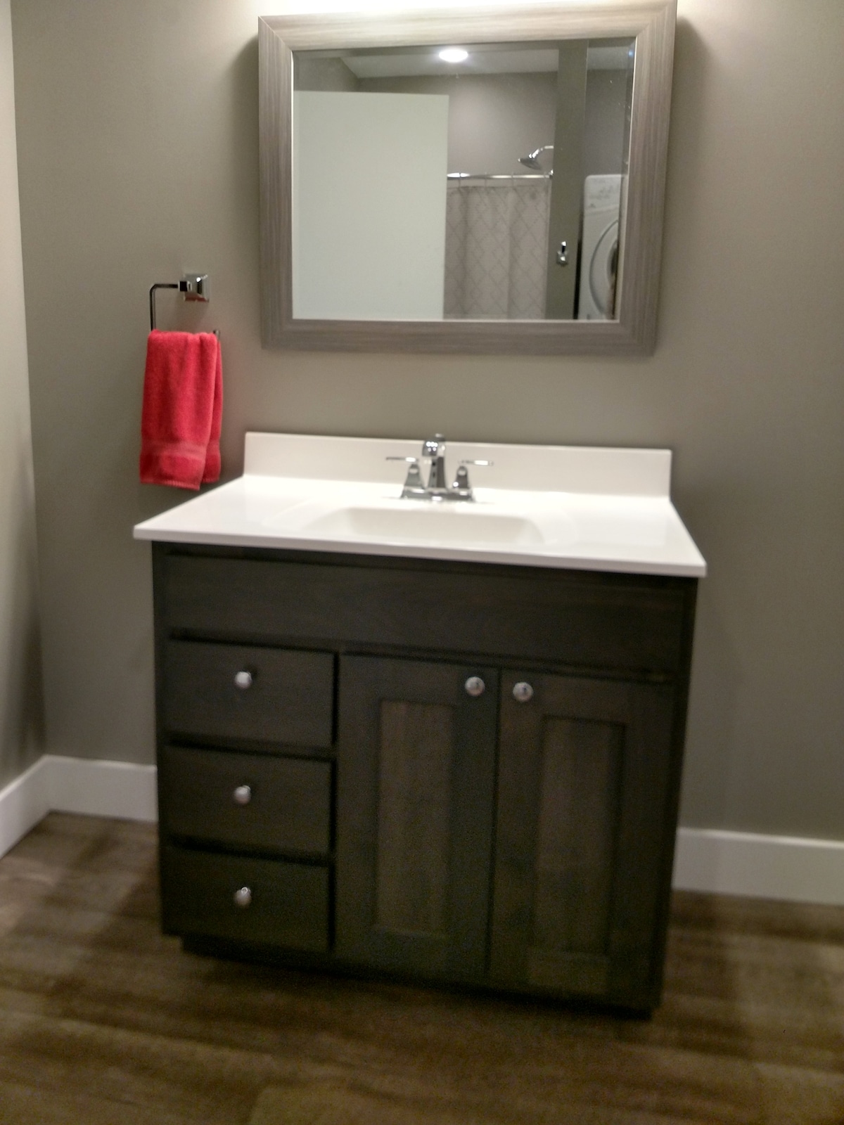 A modern bathroom sink area features a dark wood vanity with a white countertop. The vanity includes three drawers and a two-door cabinet. A mirror is mounted above the sink, with light illuminating the space. A red towel hangs from a nearby rack on the wall.