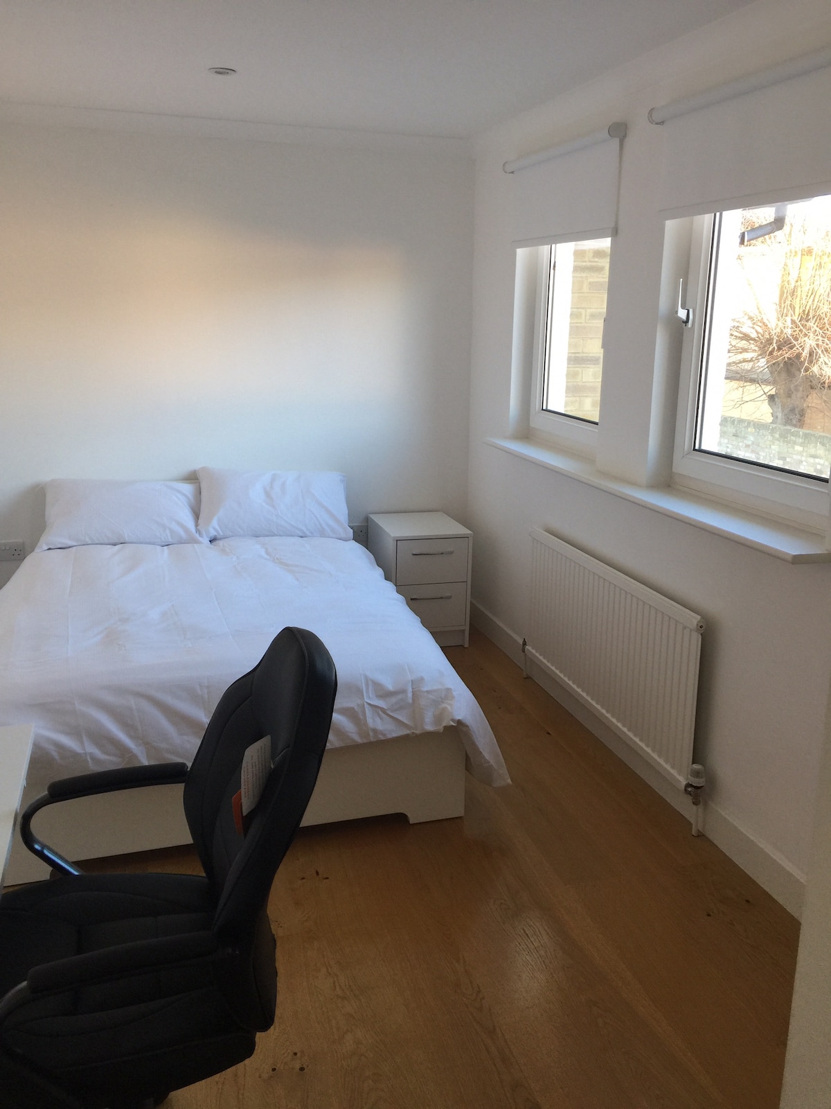 The room features a spacious double bed with white linens. A bedside table sits beside the bed, and a desk with an ergonomic chair is present. Natural light enters through two windows, illuminating the light hardwood flooring.