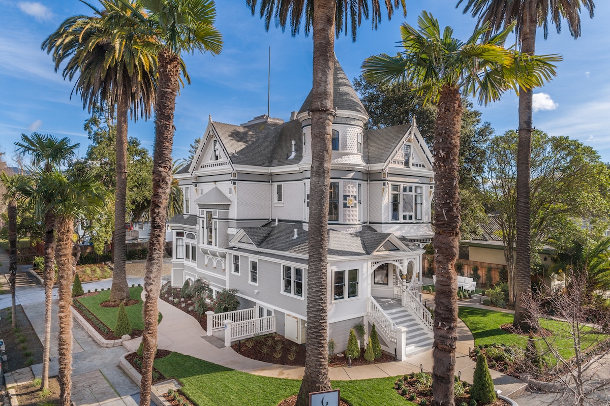 A historic white residence is framed by tall palm trees, showcasing its elegant architecture and expansive front porch. The well-maintained garden and pathway lead to the entrance, while the intricate roofline hints at the building's original design elements.