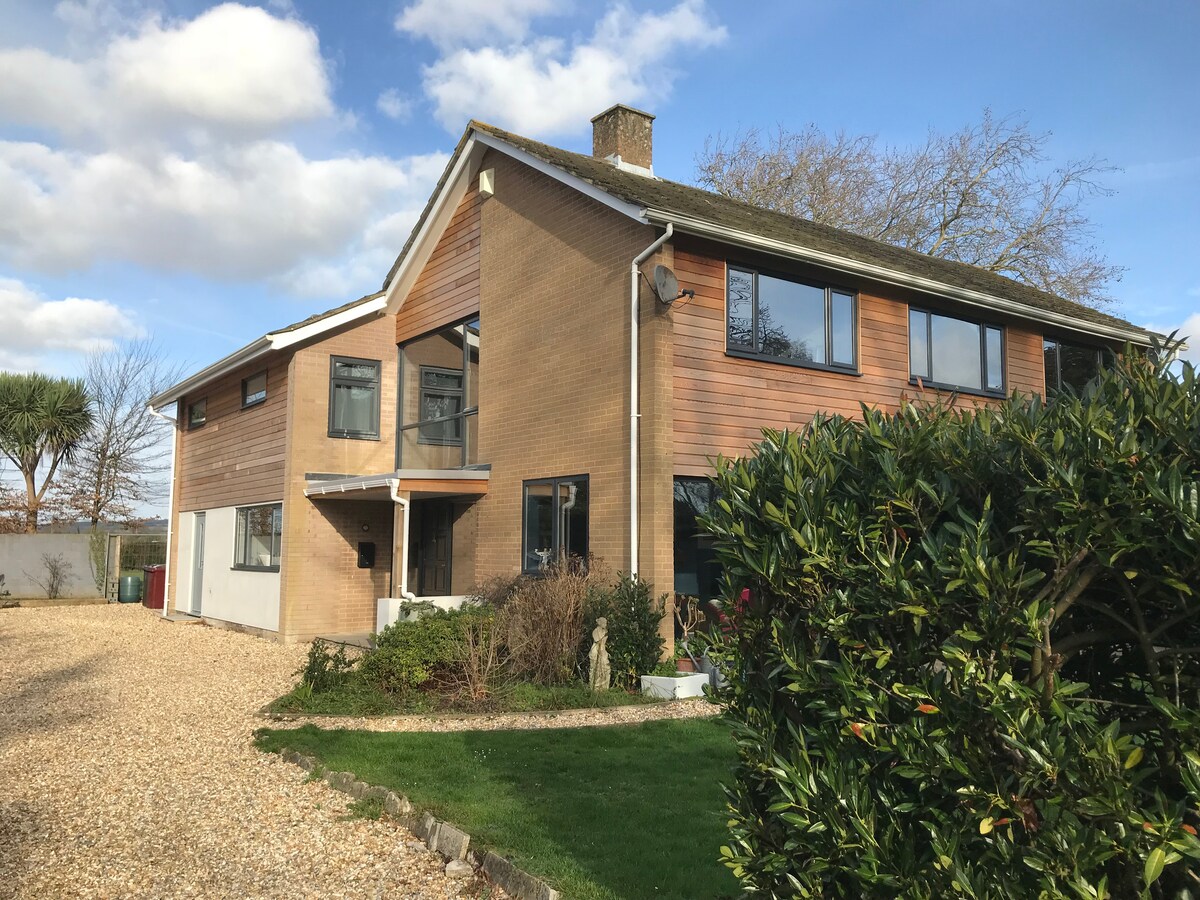 A modern independent annexe is shown, featuring a blend of brick and wood exterior. A private entrance is visible, with a landscaped garden and gravel path leading up to it. Large windows bring in natural light, and a chimney rises against a blue sky.