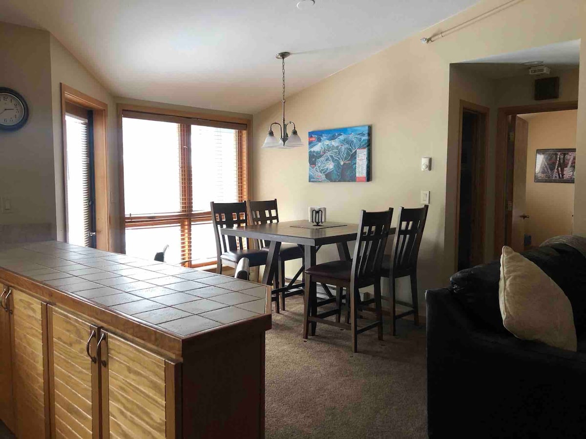 A dining area is visible, featuring a rectangular table surrounded by six chairs. Large windows allow natural light to fill the space, framed by wooden blinds. A kitchenette with tiled countertops is located in the foreground, enhancing the open layout.
