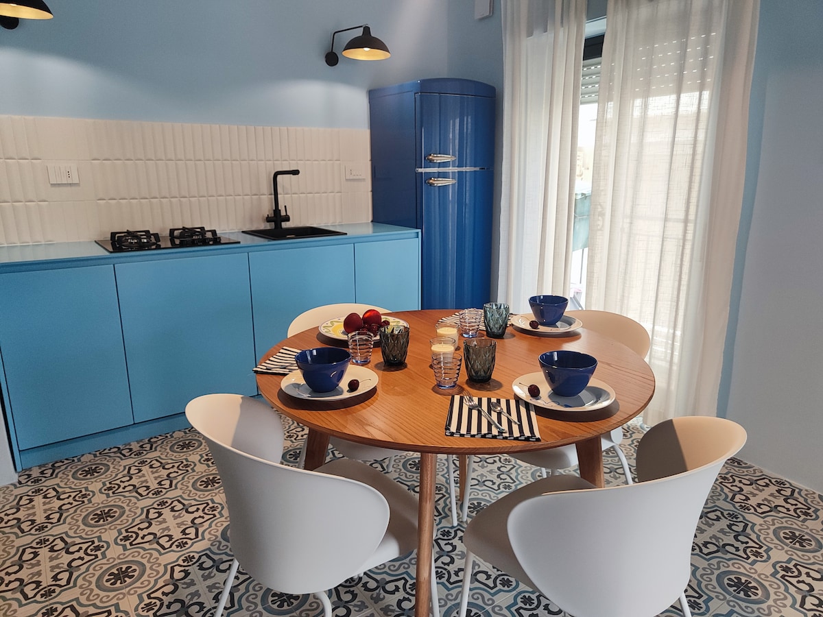 A dining area features a round wooden table set with dishes and glasses. Four white chairs surround the table, which is complemented by a blue kitchen setup in the background. A vibrant blue refrigerator adds a pop of color, with light filtering through sheer curtains.