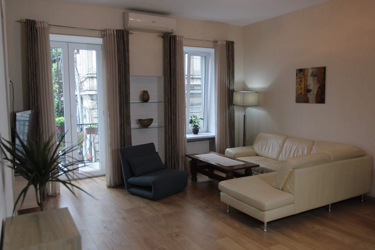A bright living area is featured, with large windows allowing natural light to fill the space. A light-colored leather sofa and a simple coffee table are present. A modern armchair sits slightly askew, and artwork adorns one wall, while a potted plant adds a touch of greenery.