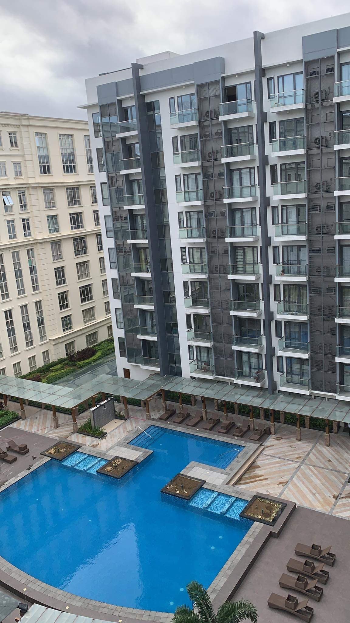 An expansive swimming pool occupies the center of the image, surrounded by multiple lounge chairs. Tall buildings frame the pool area, featuring large windows that reflect natural light. A covered walkway extends alongside the pool, enhancing accessibility to the outdoor space.