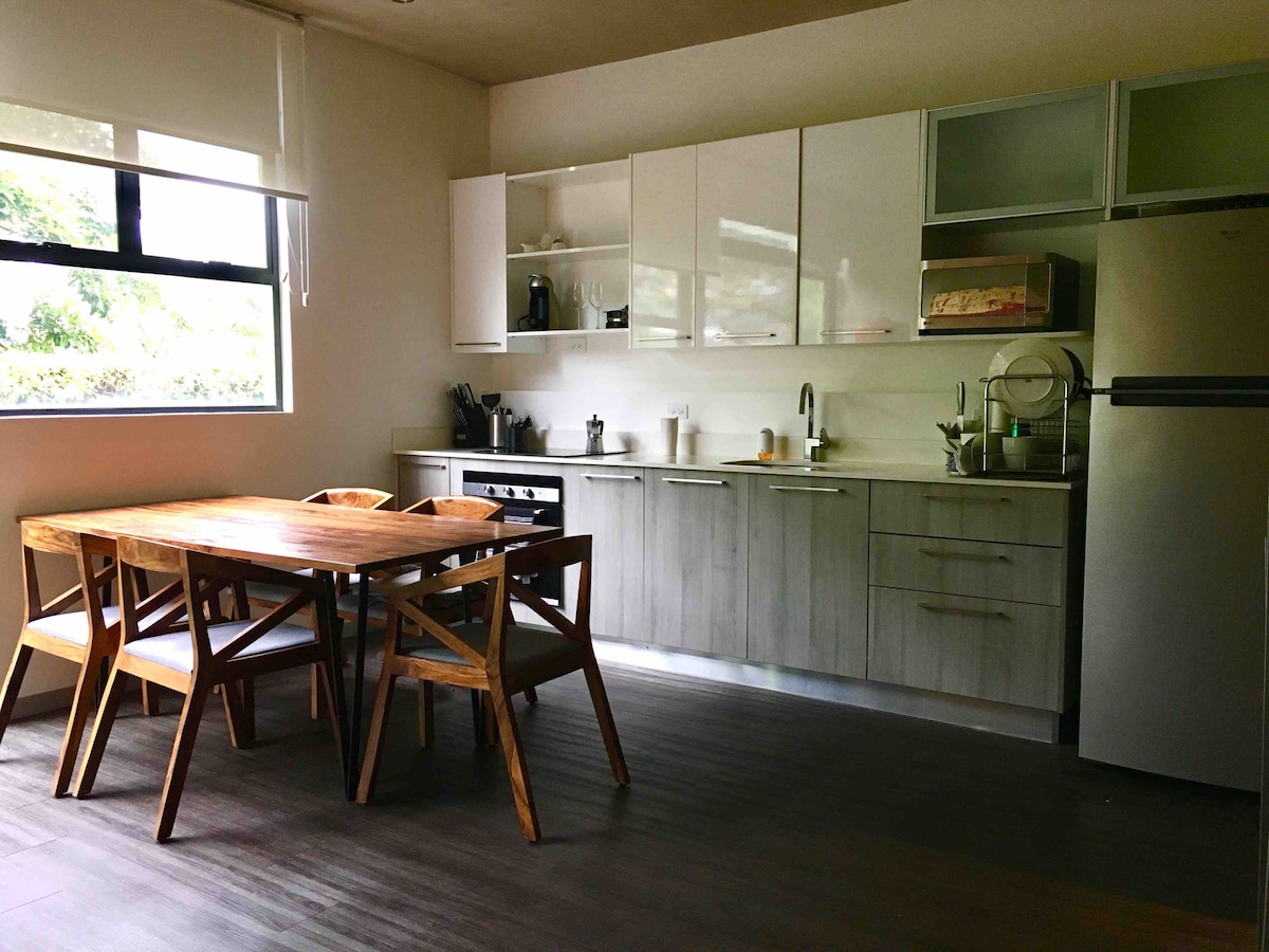 A modern kitchen features light wood cabinetry and stainless steel appliances. A wooden dining table with four chairs is positioned nearby. Natural light fills the space through a large window, creating a bright and functional cooking area.