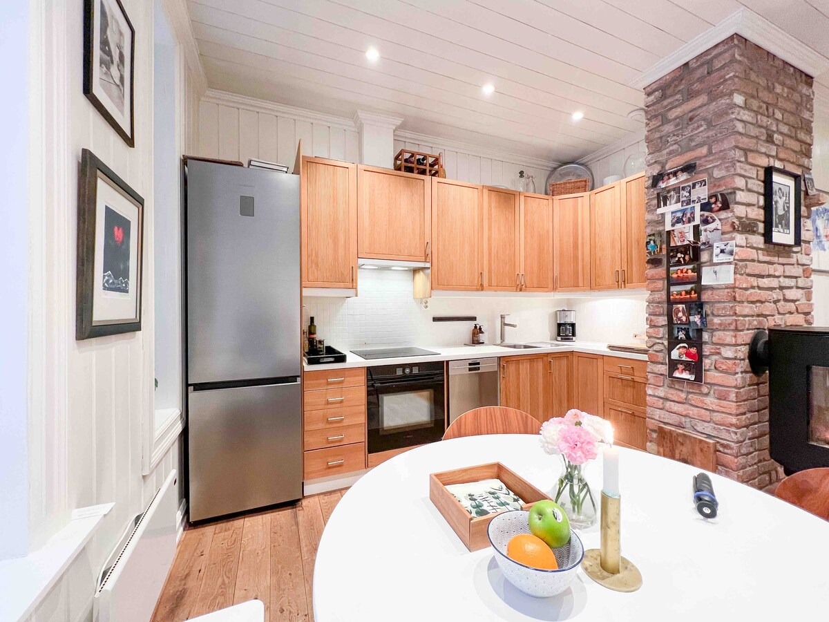 A modern kitchen is depicted, featuring wooden cabinetry and stainless steel appliances. A round dining table is set in the foreground, adorned with fresh fruits and flowers. Brick walls add texture, while ambient lighting enhances the inviting atmosphere of the space.
