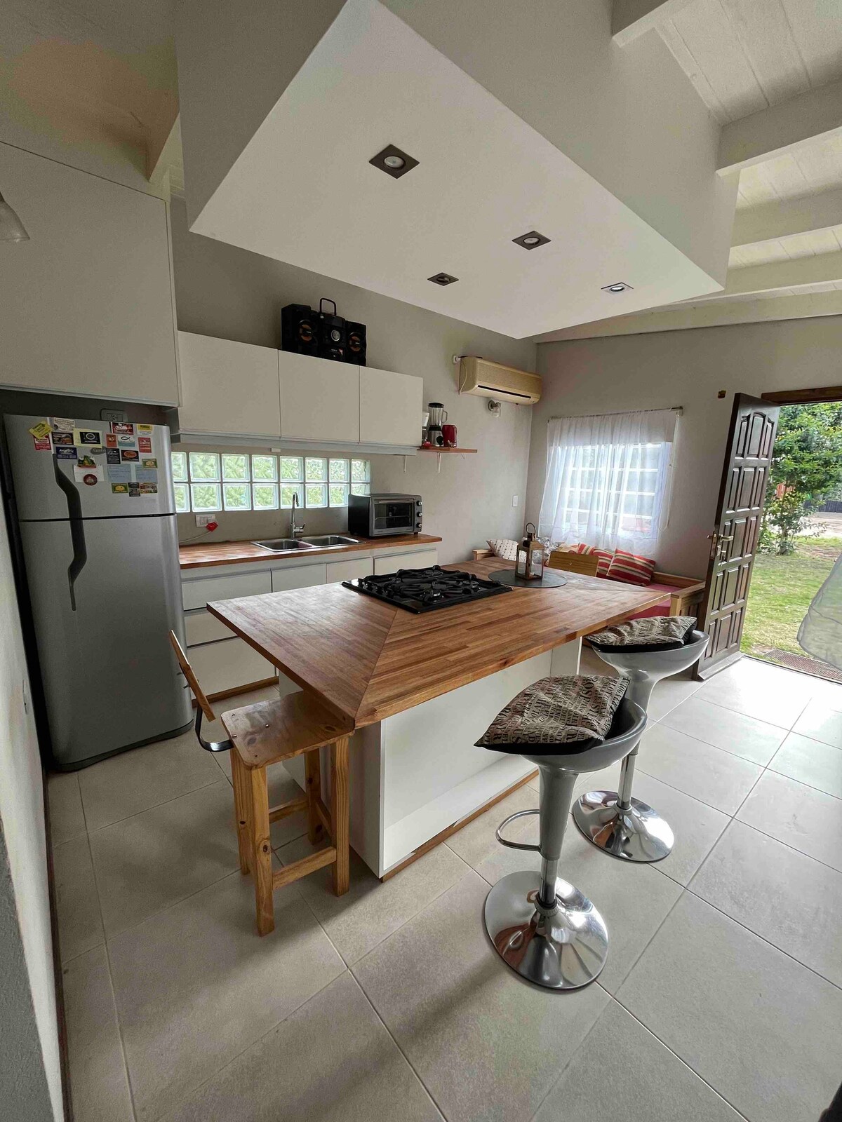 A modern kitchen is presented with a central island featuring a built-in stovetop. Sleek cabinetry and a stainless steel refrigerator are visible. Adjacent seating at the island includes four high stools. Natural light filters through a window, illuminating the functional workspace.