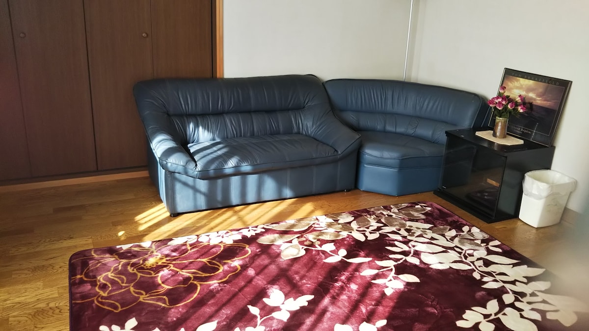 A comfortable, curved blue leather sofa is positioned in the corner, adjacent to a small black side table. A decorative rug with floral patterns in rich colors covers the wooden floor, while light from a nearby window creates dynamic shadows across the space.