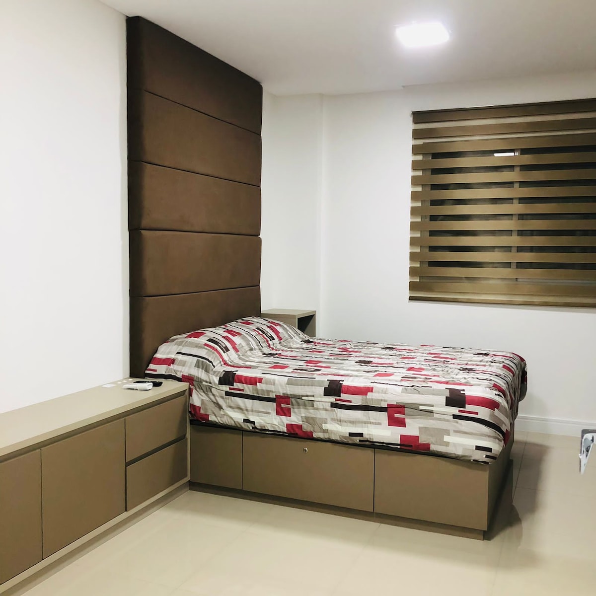 A neatly arranged sleeping area features a spacious bed dressed in a patterned duvet. A modern headboard is positioned against one wall, complemented by a side table with drawers. Soft window treatments allow filtered light to enter the room.