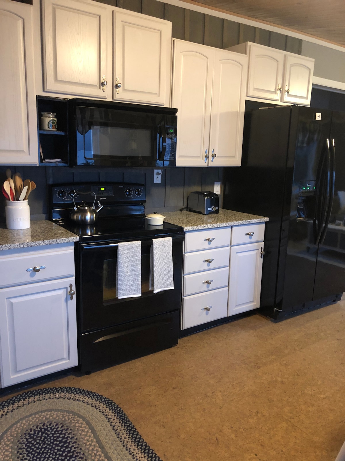 The kitchen features white cabinetry and a black stove and refrigerator. A countertop in a speckled design surrounds the appliances, complemented by practical kitchen tools on display. Soft lighting creates a welcoming ambiance, with a braided rug positioned beneath the workspace.