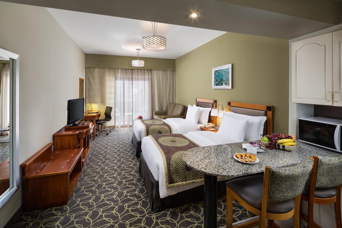 A spacious studio layout features two large beds with patterned bedding, complemented by a cozy seating area and a work desk. Natural light floods the room through large curtains, and a kitchenette is equipped with modern appliances, offering convenience for guests.