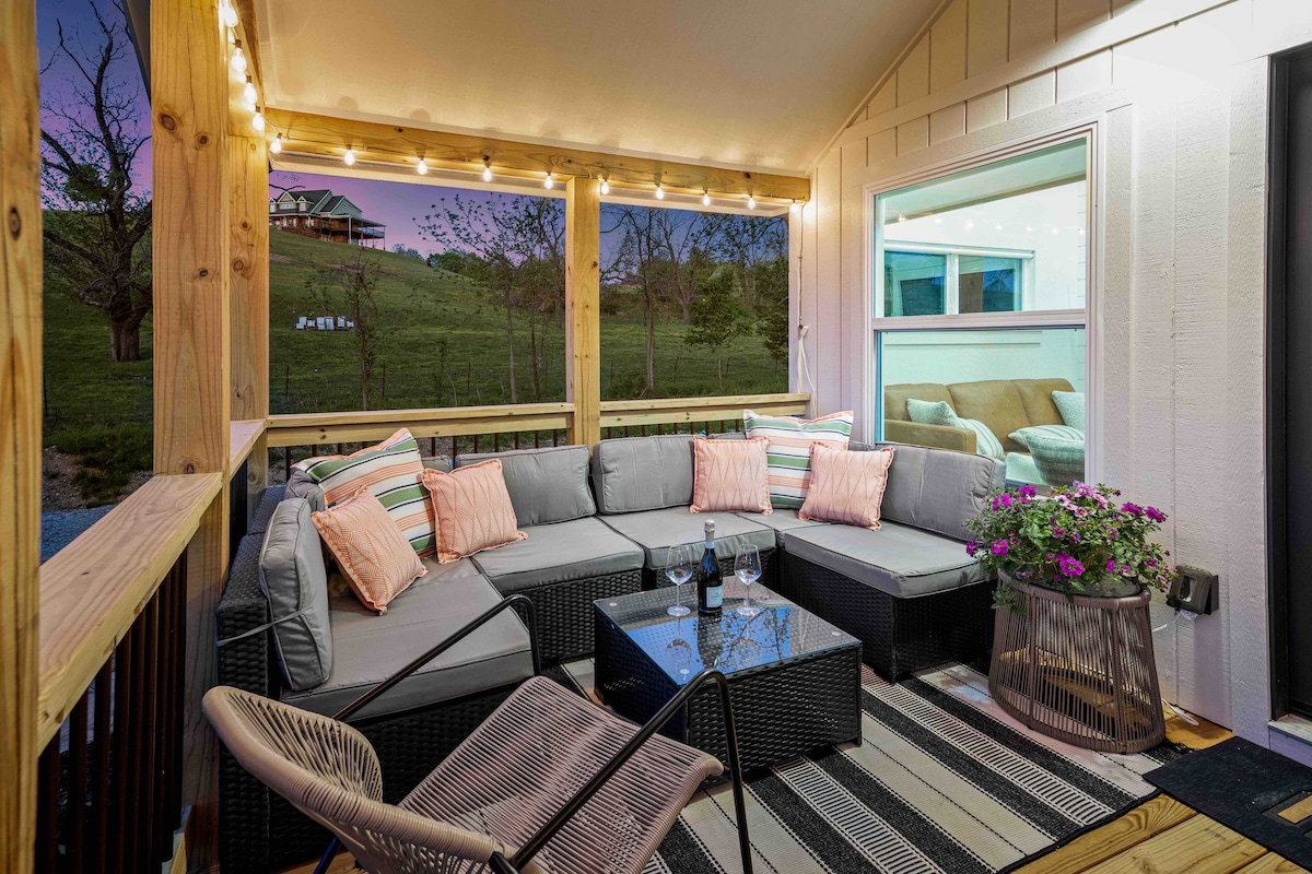 A covered porch featuring a spacious sectional sofa adorned with pillows, accompanied by a low coffee table. Soft lighting illuminates the area, creating a cozy atmosphere. A potted plant with vibrant flowers adds a touch of color. The view reveals a serene landscape beyond.