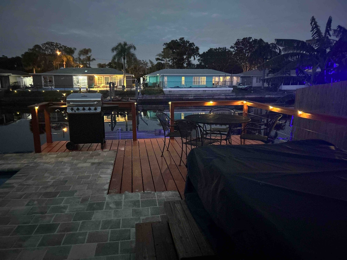 A tranquil outdoor deck overlooks the Anclote River at dusk, accented by soft lighting. A gas grill is stationed nearby, accompanied by a dining table with several chairs. In the background, charming homes are illuminated along the water's edge.
