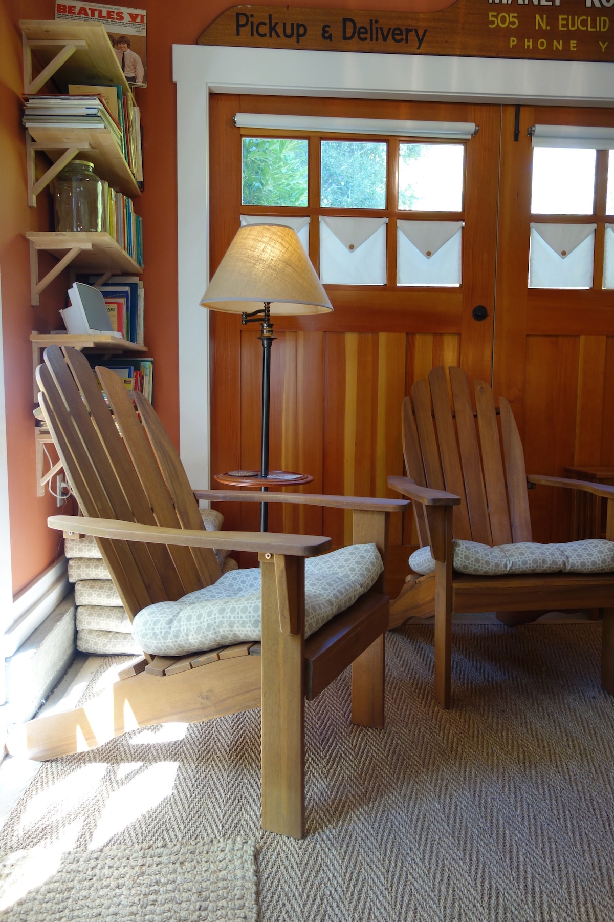 Two wooden adirondack chairs are positioned in a cozy reading nook. A soft-patterned cushion is placed on each chair. A lamp stands beside the chairs, providing warm light. The walls are painted in a warm orange hue, and a shelf lined with books is visible.