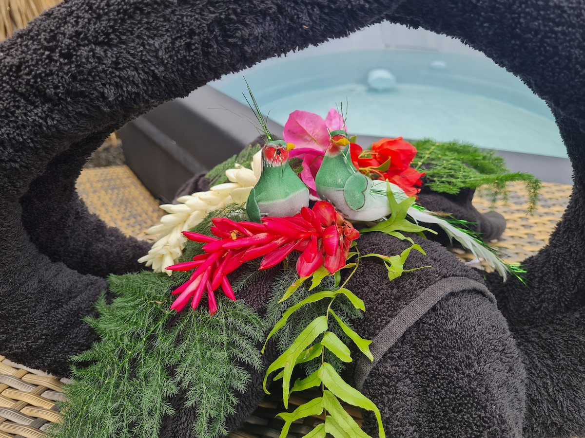 Soft, dark towels are elegantly arranged and adorned with vibrant tropical flowers and foliage. The arrangement features small decorative elements suggesting a welcoming, relaxing atmosphere, situated near a spa that can be seen in the blurred background.