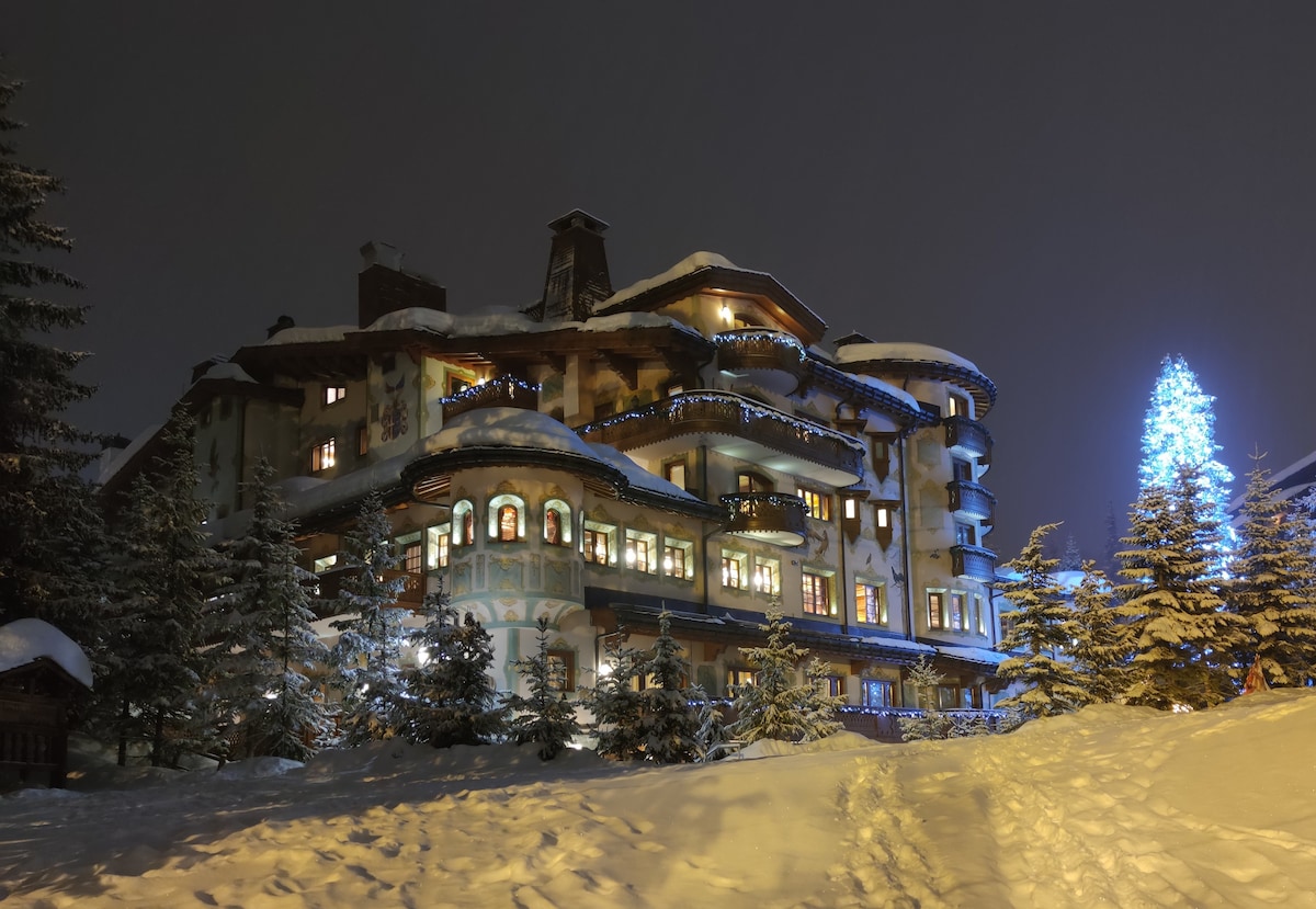 A charming chalet-style building is nestled in snow, illuminated by soft light from its windows. Surrounding evergreen trees add to the wintry scene, while a large blue tree stands brightly lit nearby. The atmosphere reflects a serene, winter evening.