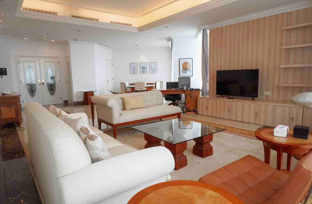 A spacious living area features a light-colored sofa set, accompanied by a wooden coffee table. A large flat-screen TV is mounted on a wooden cabinet. The room is illuminated by natural light through large windows, enhancing the minimalist and elegant design.