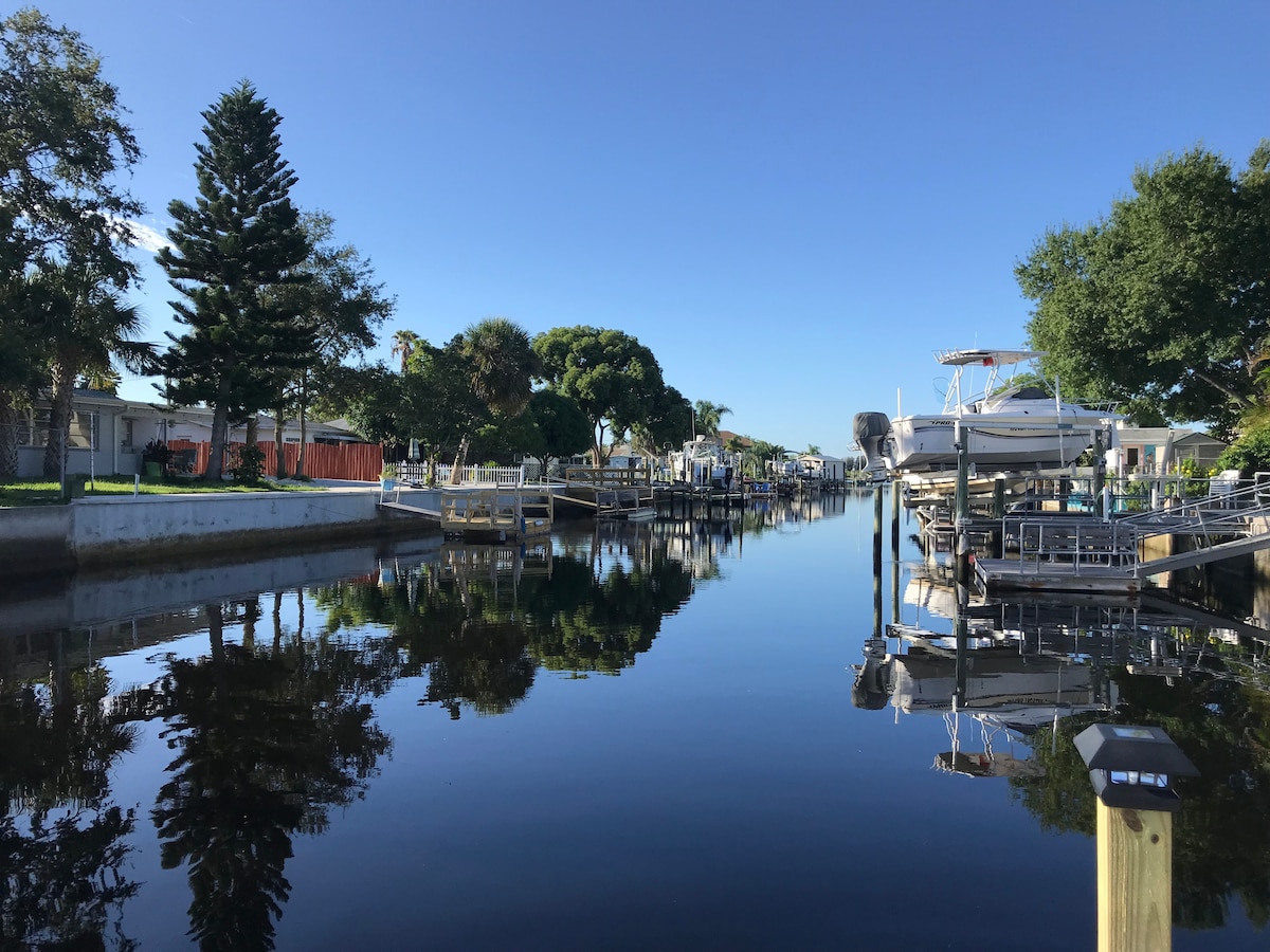 A tranquil canal scene is captured, reflecting lush greenery and residential areas along the waterway. Boats are docked on both sides, and a clear blue sky is mirrored in the calm water, creating a serene atmosphere enhanced by gentle natural surroundings.