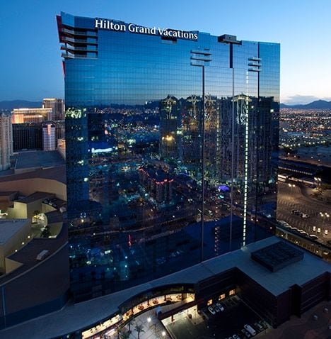 The hotel stands tall against the evening sky, its reflective glass facade capturing the surrounding city lights. The modern architecture is complemented by a lower-level area showcasing entrances and walkways, blending seamlessly with the vibrant atmosphere of the Las Vegas Strip.