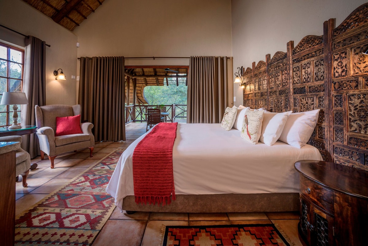 A spacious bedroom features a king-sized bed adorned with white linens and decorative pillows. Natural light enters through large windows, revealing a serene view of the outdoors. Two comfortable chairs and a wooden table complement the space, while intricate wooden screens add aesthetic detail.