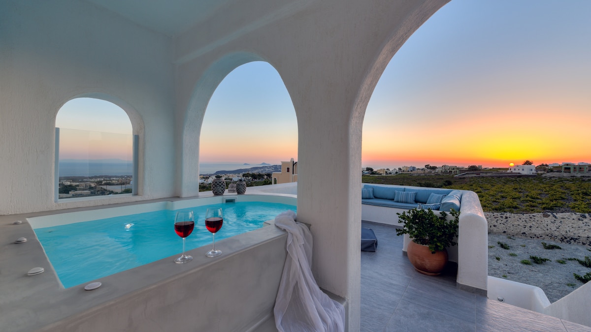 A serene balcony features a private jacuzzi overlooking a scenic sunset. Two glasses of red wine are placed on the ledge beside a soft towel. Comfortable seating is visible in the background, framed by architectural arches that enhance the view of the Aegean Sea.