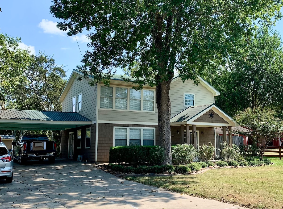 The exterior of a two-story house is shown, featuring a light-colored facade with large windows. A carport is visible on the left, and a well-maintained lawn surrounds the property, providing shade from a large tree in the front yard. A welcoming entrance is highlighted.