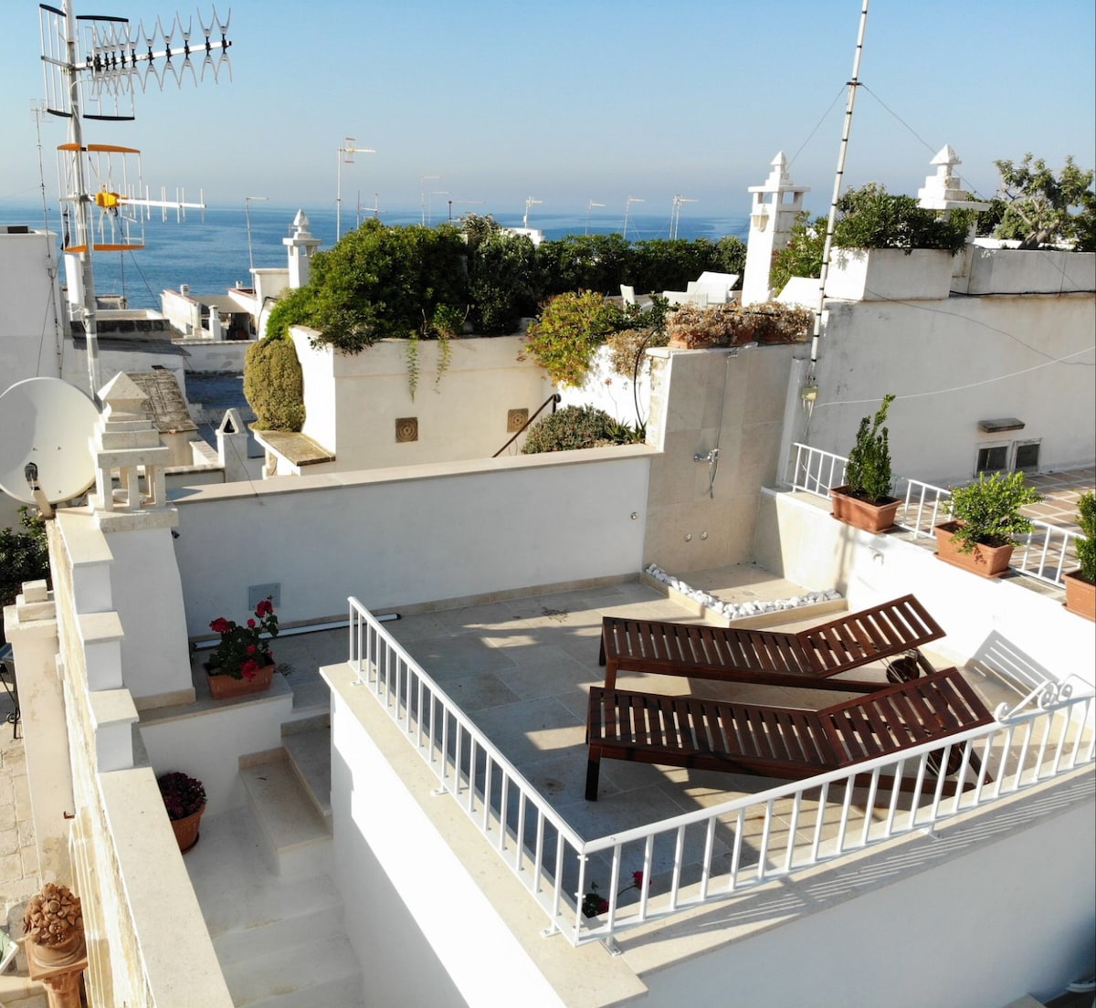 A sunlit terrace features two lounge chairs and potted plants against a backdrop of the sea. The white railing provides safety while offering unobstructed views. The surrounding buildings are painted in light hues, contributing to the serene atmosphere of the outdoor space.