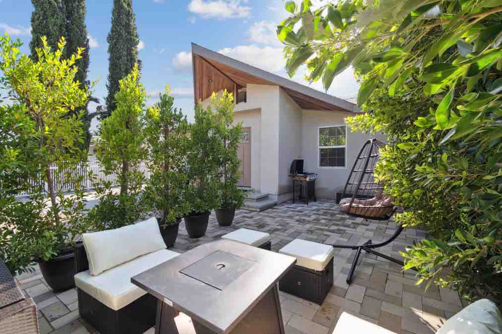 An outdoor patio space is shown, featuring comfortable seating arrangements with white cushions surrounding a square fire table. Lush green plants create a private ambiance, while a swinging chair adds a cozy touch. The patio is paved with gray stone, complementing the modern architecture of the home.