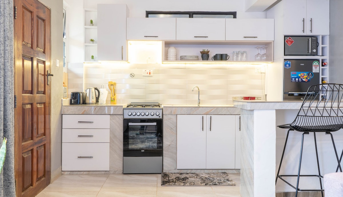 A modern kitchen area features sleek cabinetry and a minimalist design. Stainless steel appliances are included, along with a countertop that provides space for food preparation. A small area rug adds texture, while a black bar stool complements the kitchen's contemporary aesthetic.