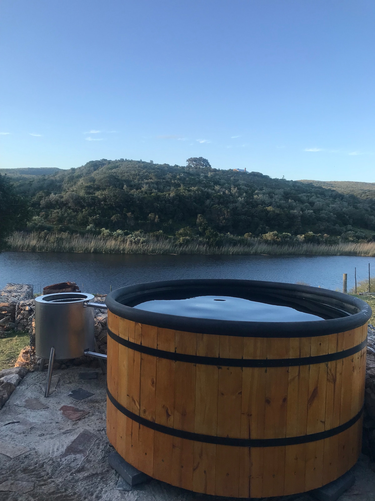 A wooden hot tub is situated outdoors, with a serene view of the river and the surrounding green hills. The calm water reflects the clear sky, creating a peaceful atmosphere. Nearby, a metal container is visible, suggesting potential use for heating purposes.