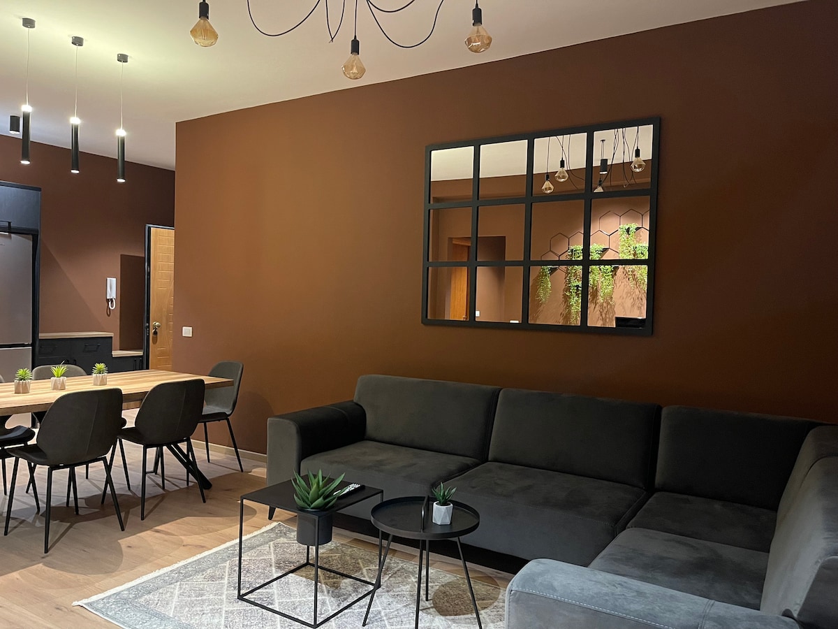 A spacious living area features a large dark gray sectional sofa, accompanied by a black coffee table and potted plants. A wooden dining table sits nearby, surrounded by black chairs. The warm brown walls are complemented by a mirrored wall decor, enhancing the room's modern aesthetic.