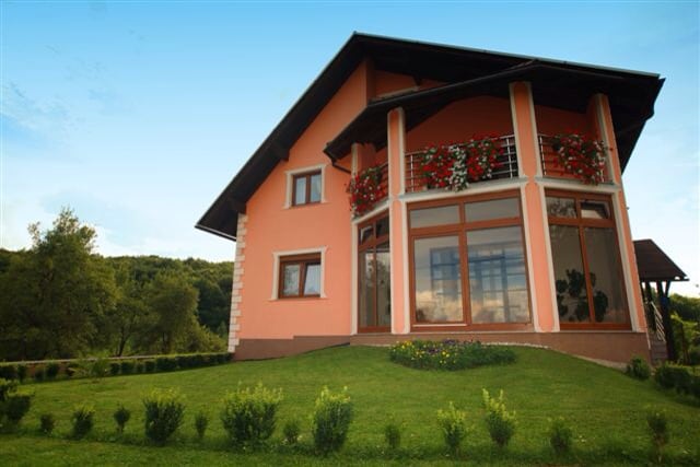 A charming two-story house is presented, featuring large glass windows that allow natural light to illuminate the interior. The exterior is painted in soft shades of pink, complemented by vibrant flowers in window boxes. A well-maintained lawn with shrubs frames the residence.
