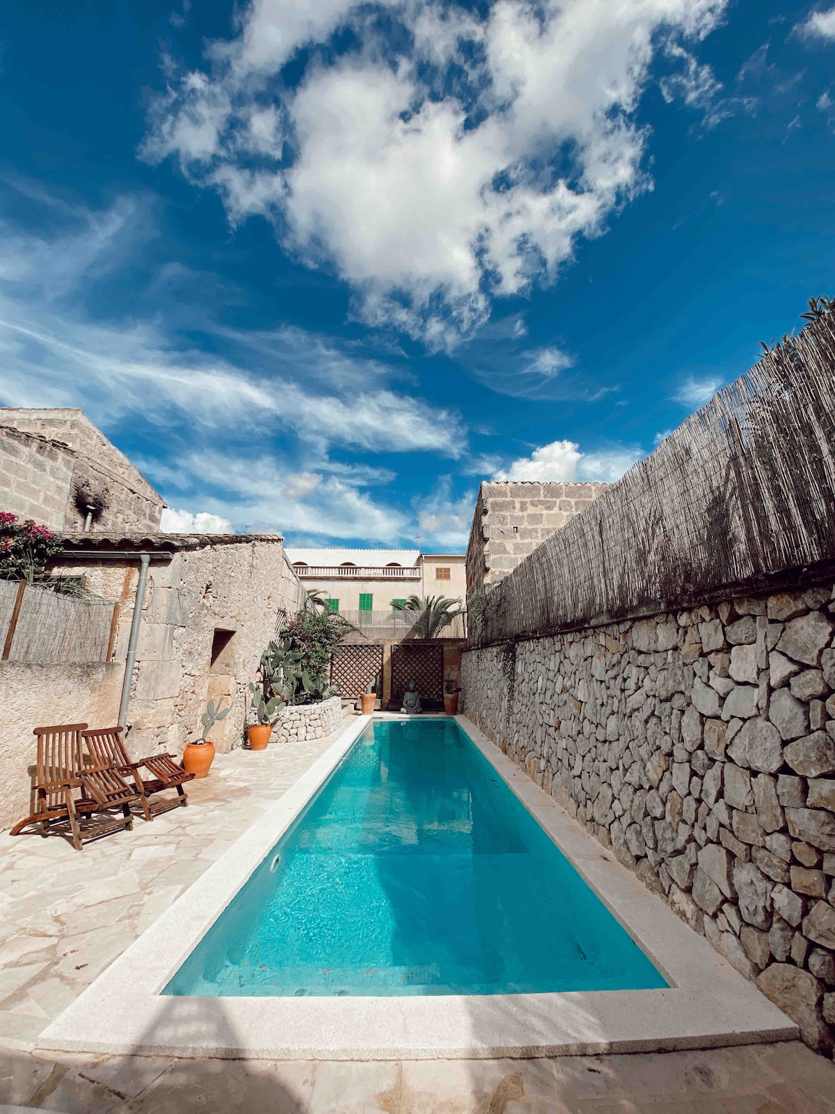 A narrow swimming pool, bordered by natural stone walls, stretches under a bright blue sky with scattered clouds. Wooden lounge chairs and terracotta planters are placed along the poolside, creating an inviting outdoor space for relaxation.