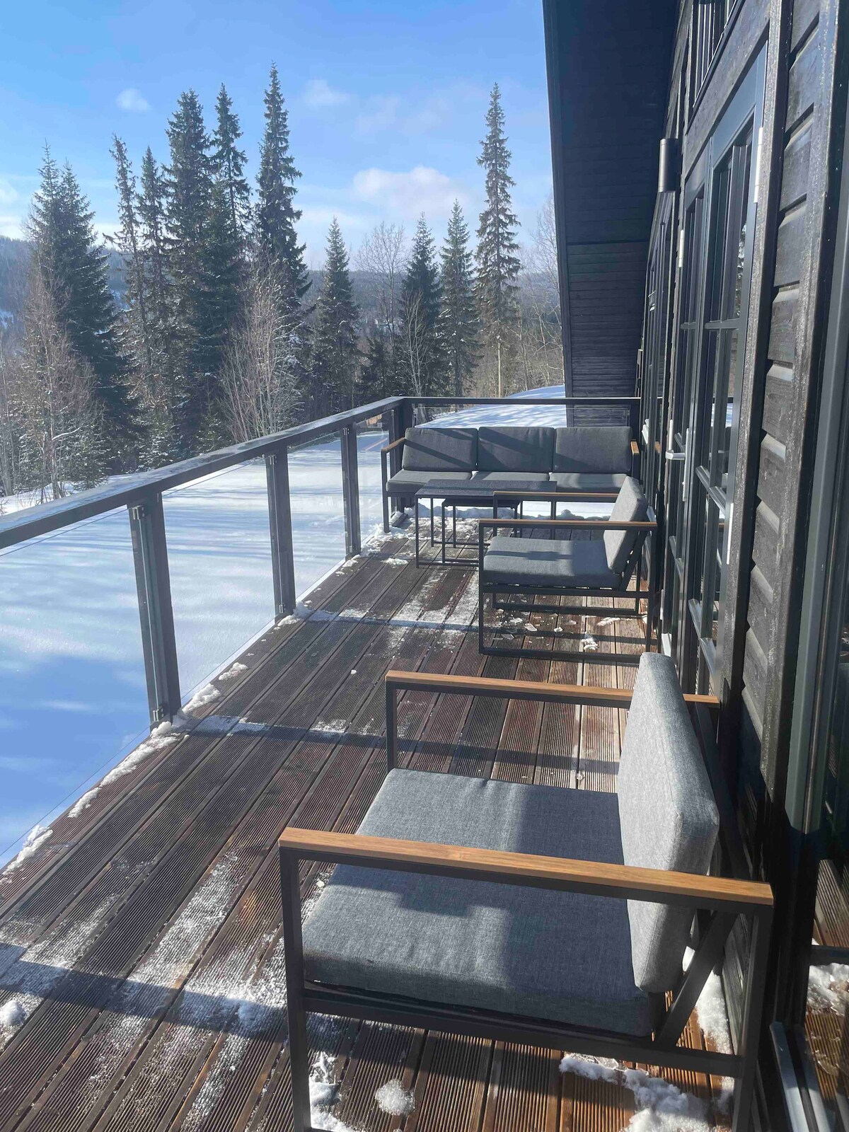 A wooden balcony extends alongside the property, featuring several comfortable seating options. A clear railing provides a scenic view of the surrounding forest and snow-covered landscape. The area is illuminated by natural light, creating a serene outdoor space.
