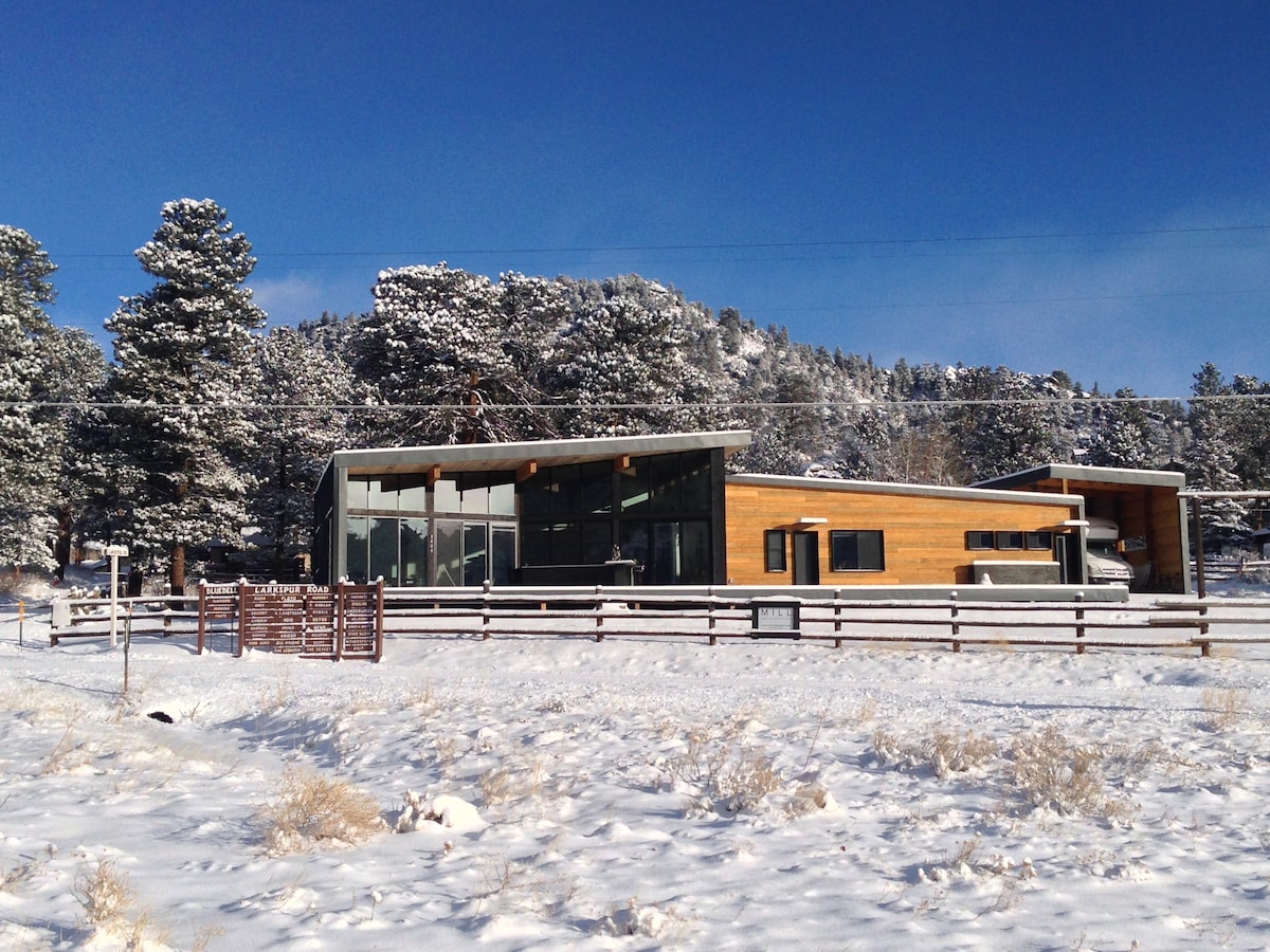 A modern cabin sits amidst a snowy landscape, framed by tall evergreen trees. Large glass windows enhance the structure, reflecting the bright blue sky. Snow blankets the ground, and a wooden fence outlines the property, creating a serene winter scene.