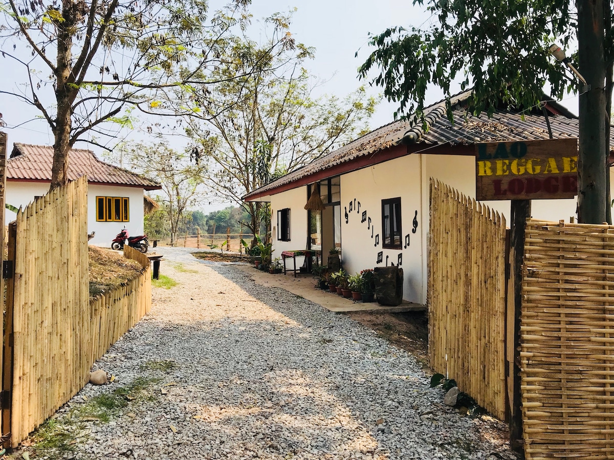 A dirt pathway leads to the two structures of Le Reggae Lodge. The entrance features a wooden fence, while the buildings are framed by trees and open space. A small seating area is visible to the side, complementing the relaxed environment.