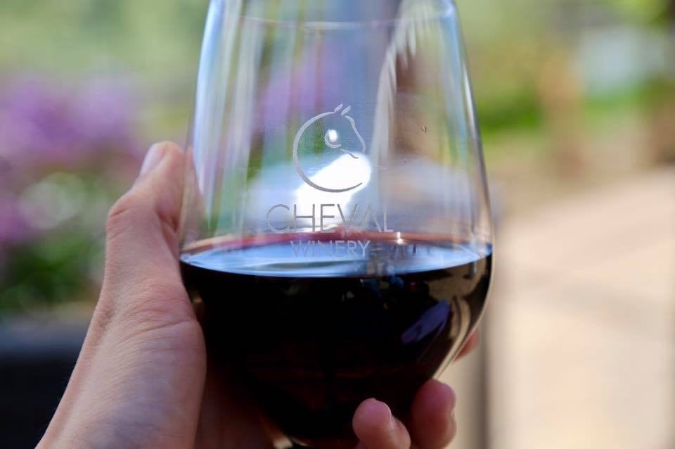 A hand holds a clear wine glass featuring the etched logo of Cheval Winery. The glass is filled with deep red wine, and a blurred garden scene with greenery and flowers can be seen in the background.