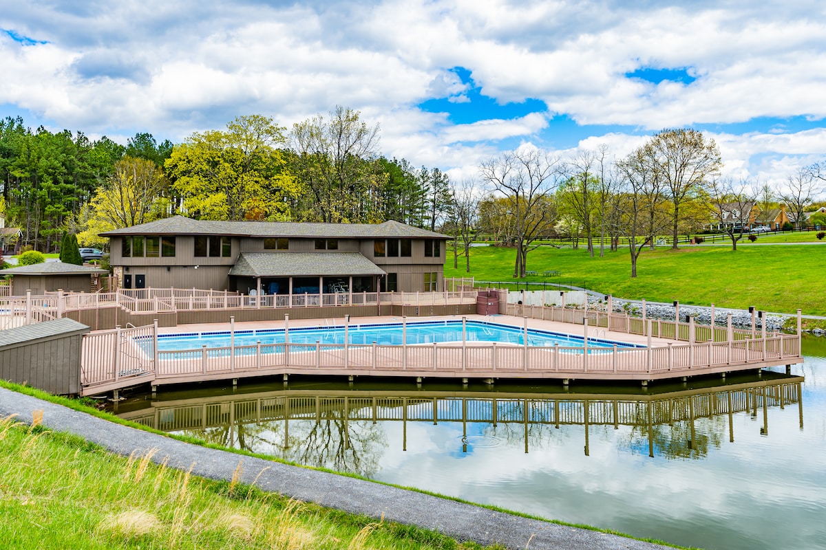 A community sports center is seen featuring a large outdoor pool surrounded by wooden decking. The pool's reflection can be observed on the calm water, creating a serene atmosphere. Lush green grass and trees are visible in the background, under a partly cloudy sky.
