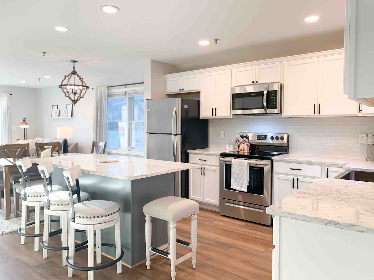 The open kitchen features modern stainless steel appliances, including a refrigerator and stove. A spacious island with Quartz countertops provides seating for four, complemented by white bar stools. The dining area is visible, showcasing a dining table set for six, with natural light flowing in from nearby windows.