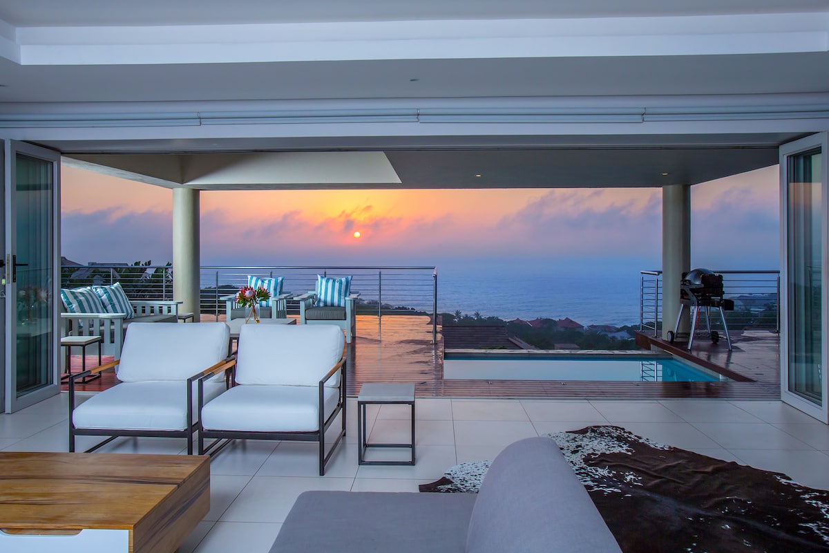 An open-plan living space is showcased with comfortable seating and a view of a sunset over the sea. Glass doors provide access to an entertainment deck and splash pool, while an outdoor dining area is visible in the background, framed by soft lighting.