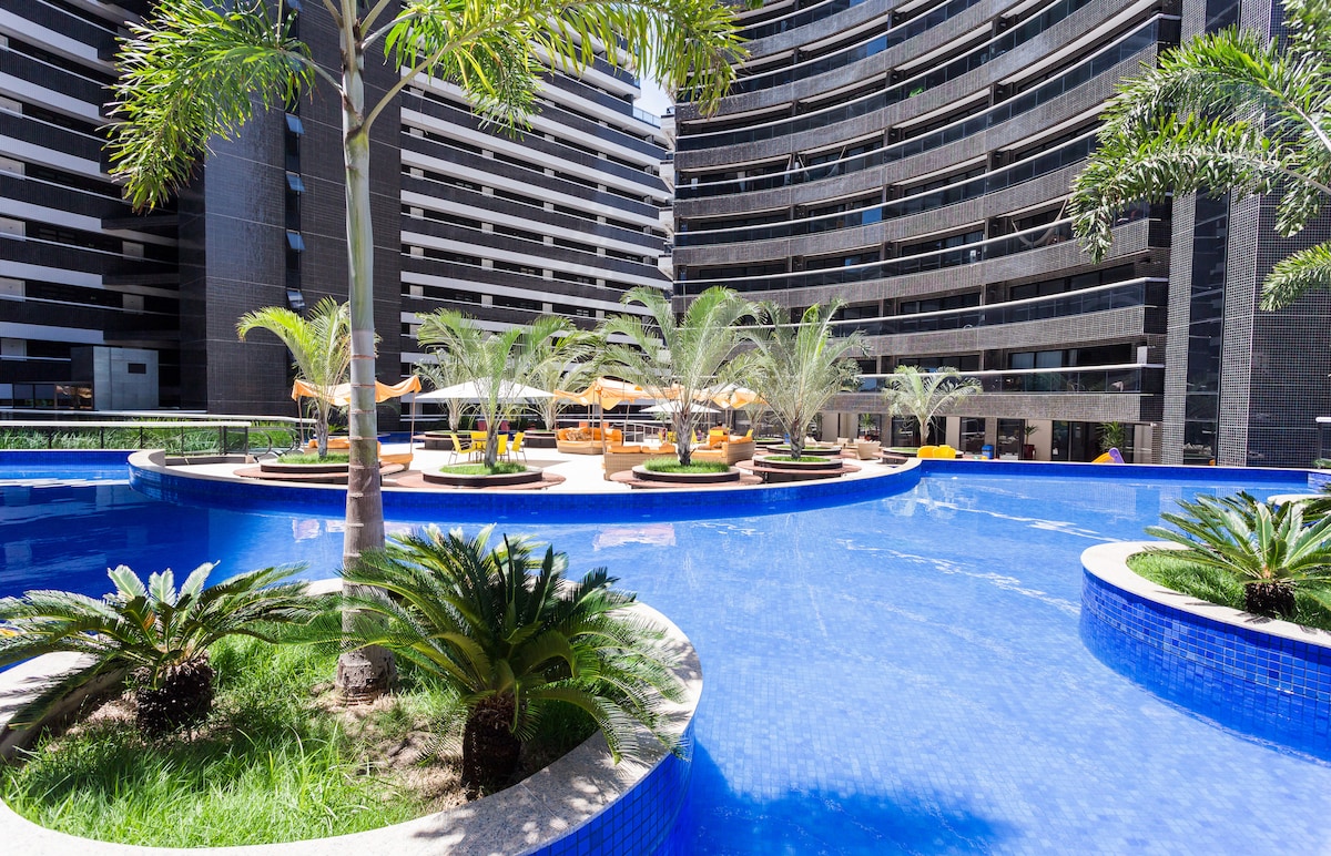 A spacious pool area features a vibrant blue surface surrounded by palm trees and comfortable lounge chairs. Umbrellas provide shade, and the modern building frames the scene with its unique architectural design, creating an inviting space for relaxation.
