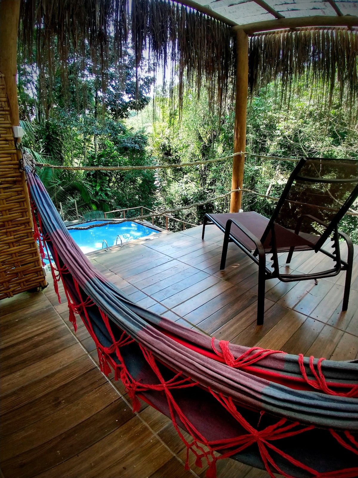 A wooden deck features a comfortable lounge chair positioned next to a hammock with bright red accents. Lush greenery surrounds the space, providing a serene escape. A glimpse of a small blue pool is visible just beyond the deck, enhancing the natural atmosphere.