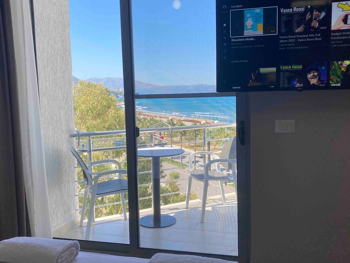 A view from the balcony showcases the sea and coastal area through large glass doors. A small round table and two chairs are positioned on the balcony, providing a relaxing outdoor space. A television mounted on the wall displays various options, completing the inviting scene.