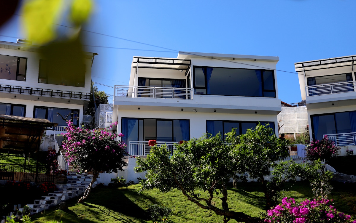 A modern structure is situated on a hillside, showcasing expansive glass windows and a balcony. Lush greenery, including blooming purple flowers and well-maintained lawns, surrounds the property, creating an engaging contrast with the clear blue sky.