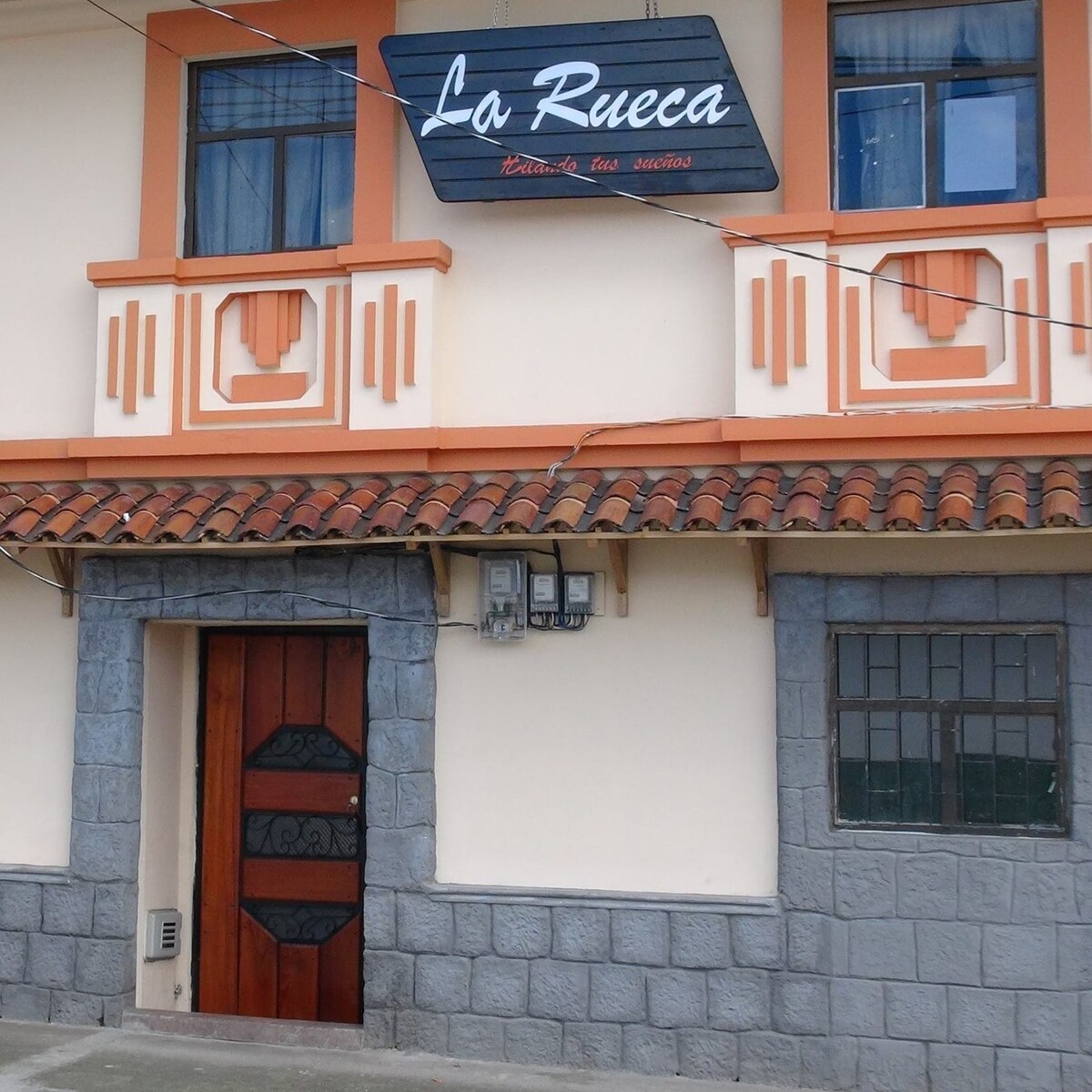 The exterior view of Casa de Hospedaje La Rueca showcases a charming facade with a welcoming wooden door framed by stone details. Above, the prominent sign displays the name, complemented by two windows adorned with simple, straight lines.